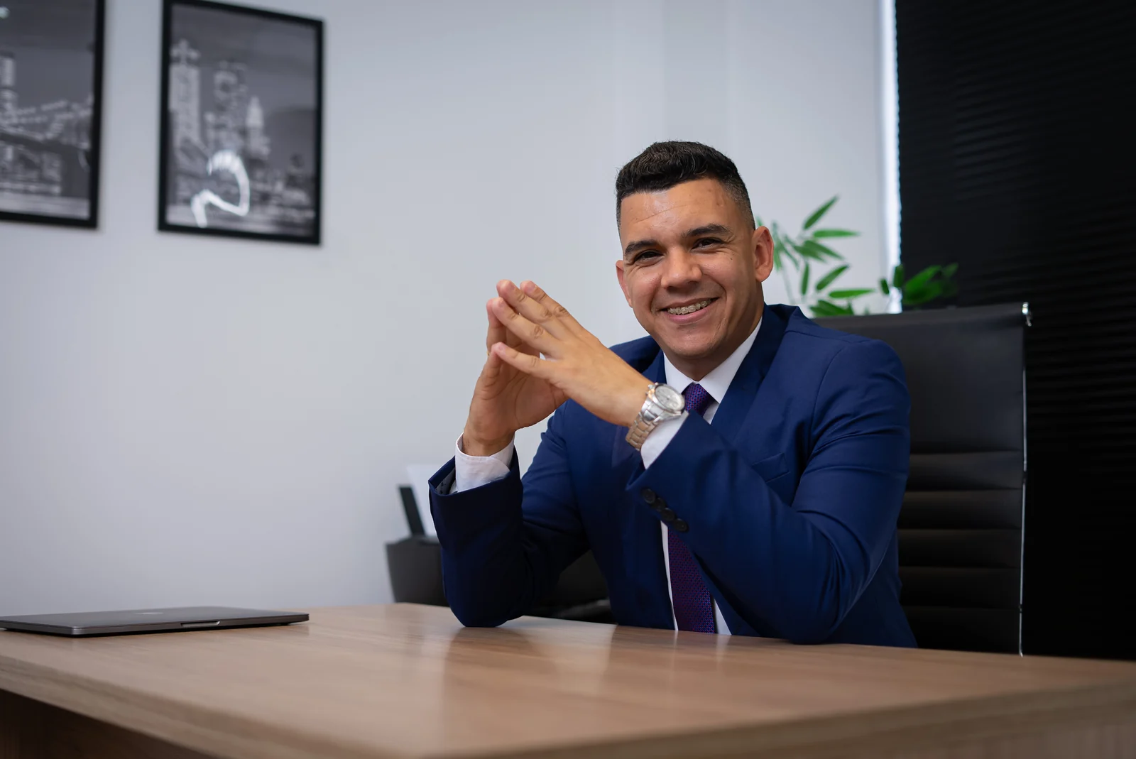 Helder Nangino sorrindo, de terno azul, sentado à mesa do escritório